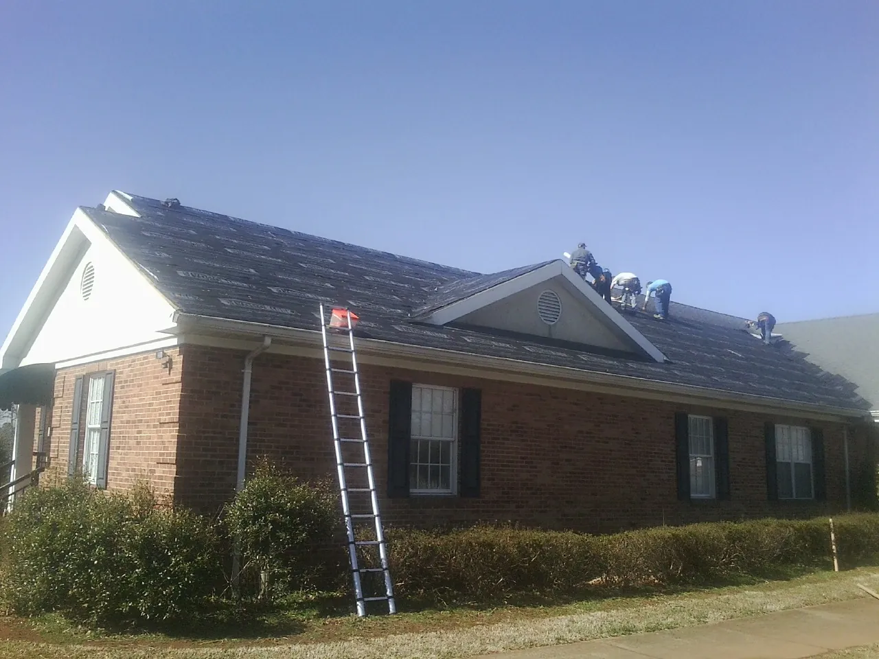 Crew tearing shingles off a brick ranch roof with a ladder set — Emergency Roof Repair work by our crew in Roswell